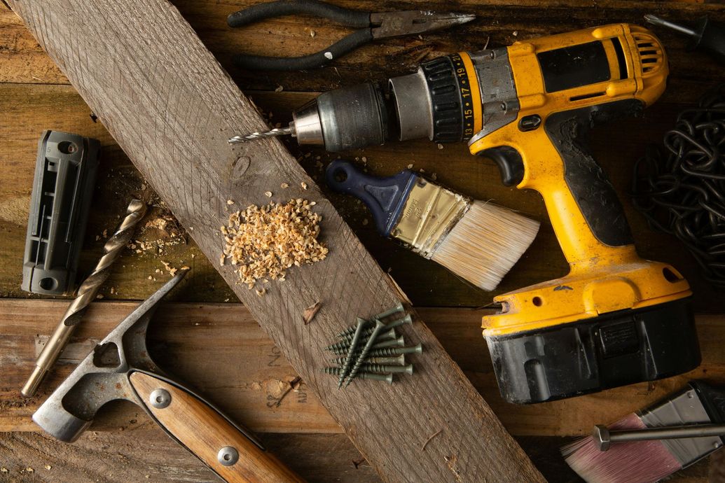 A yellow power drill, hammer, paintbrush, and hardware scattered on a wooden plank covered in sawdust.