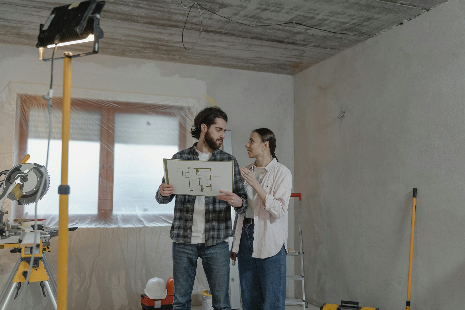 Two people in a room under renovation stand together and discuss architectural floor plans held in their hands.