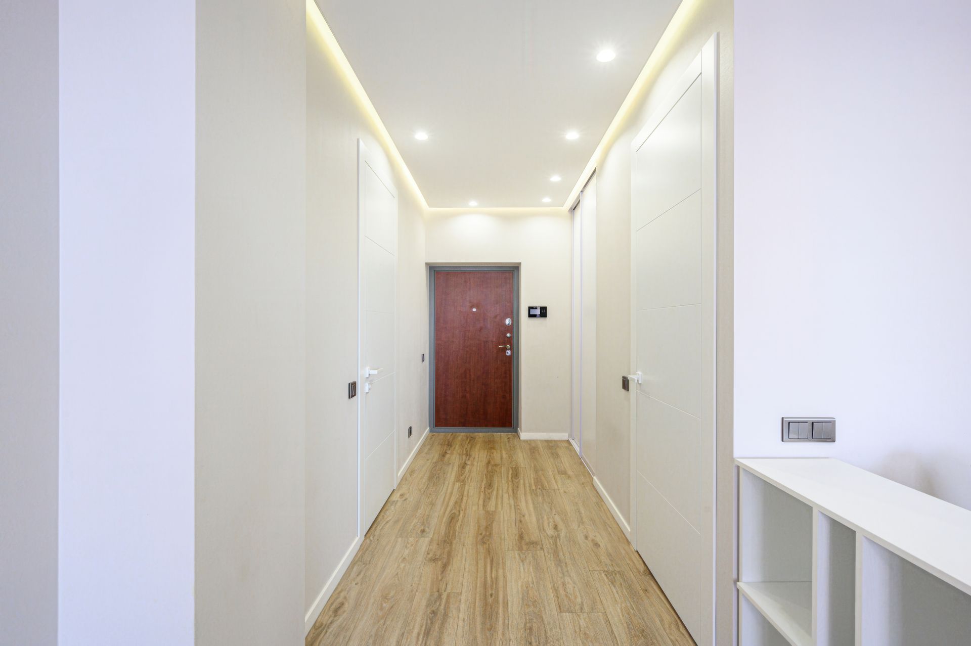 Narrow hallway with light wood-look flooring, white walls, doors, and a red door at the end.