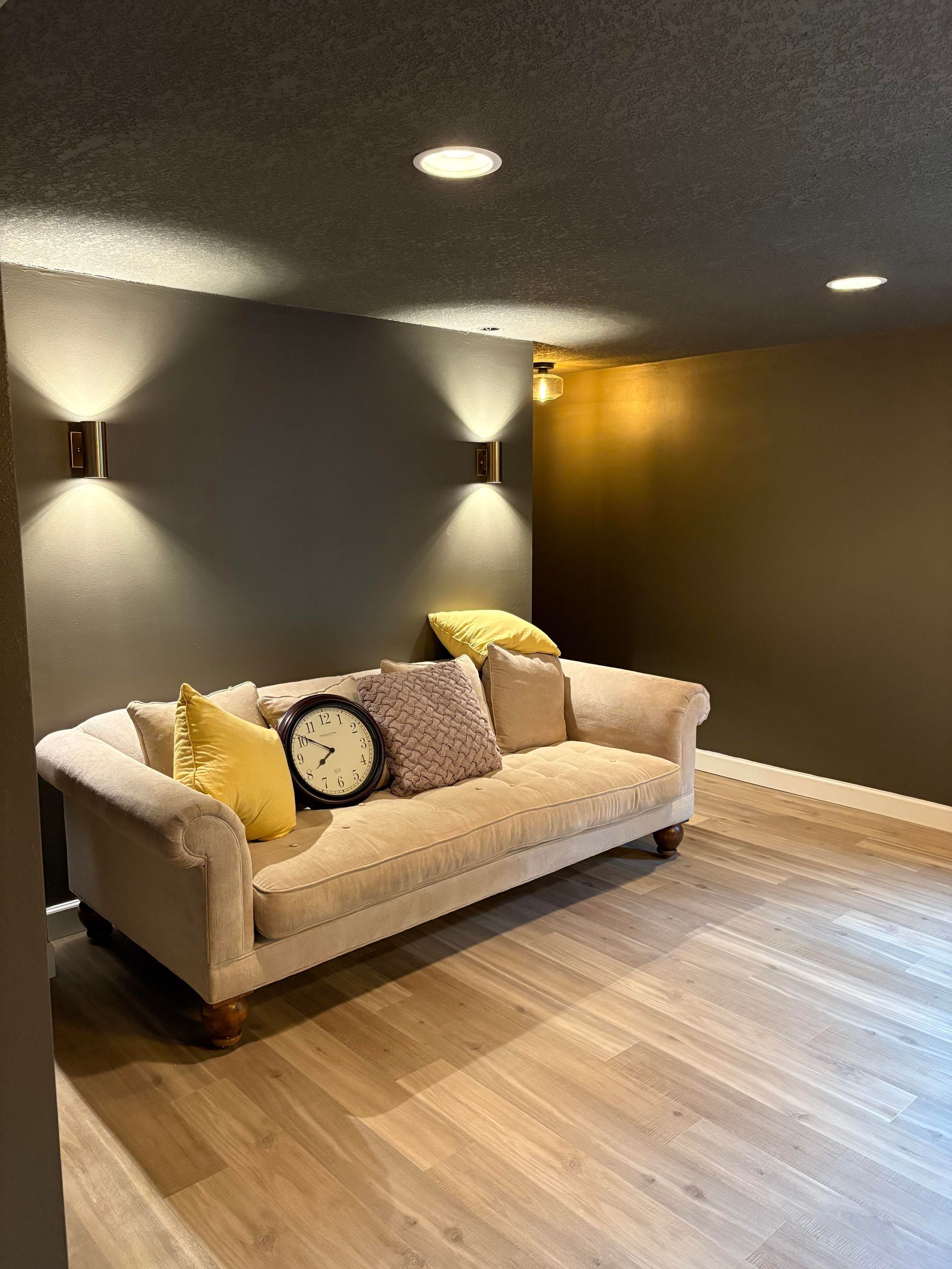 Beige sofa with pillows, lit by wall sconces, in room with wood floor and dark gray walls.