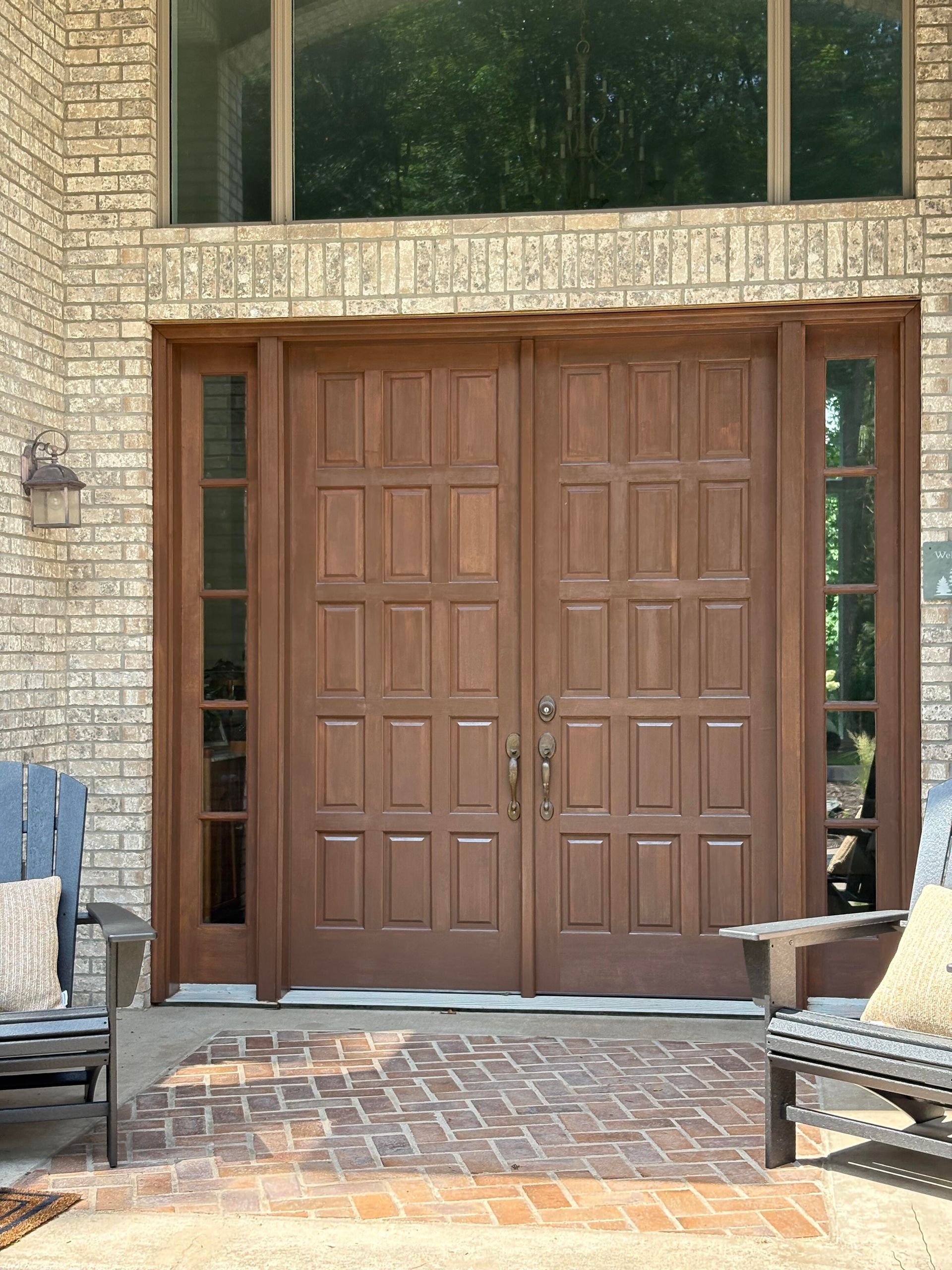 Wooden double doors with side panels set into a brick facade, flanked by chairs, leading to a home's entrance.