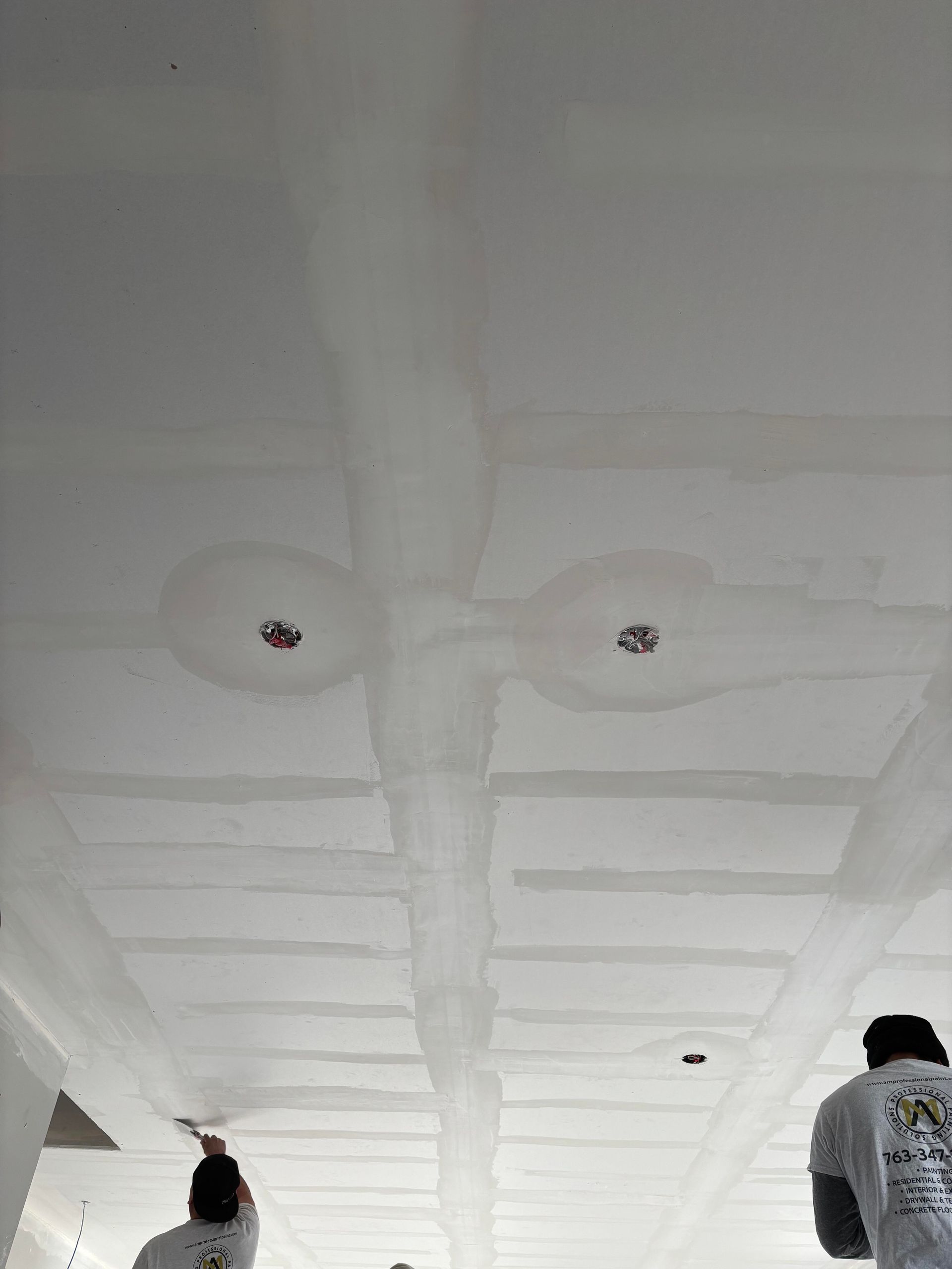 Workers applying compound to a drywall ceiling, covering seams and light fixtures.