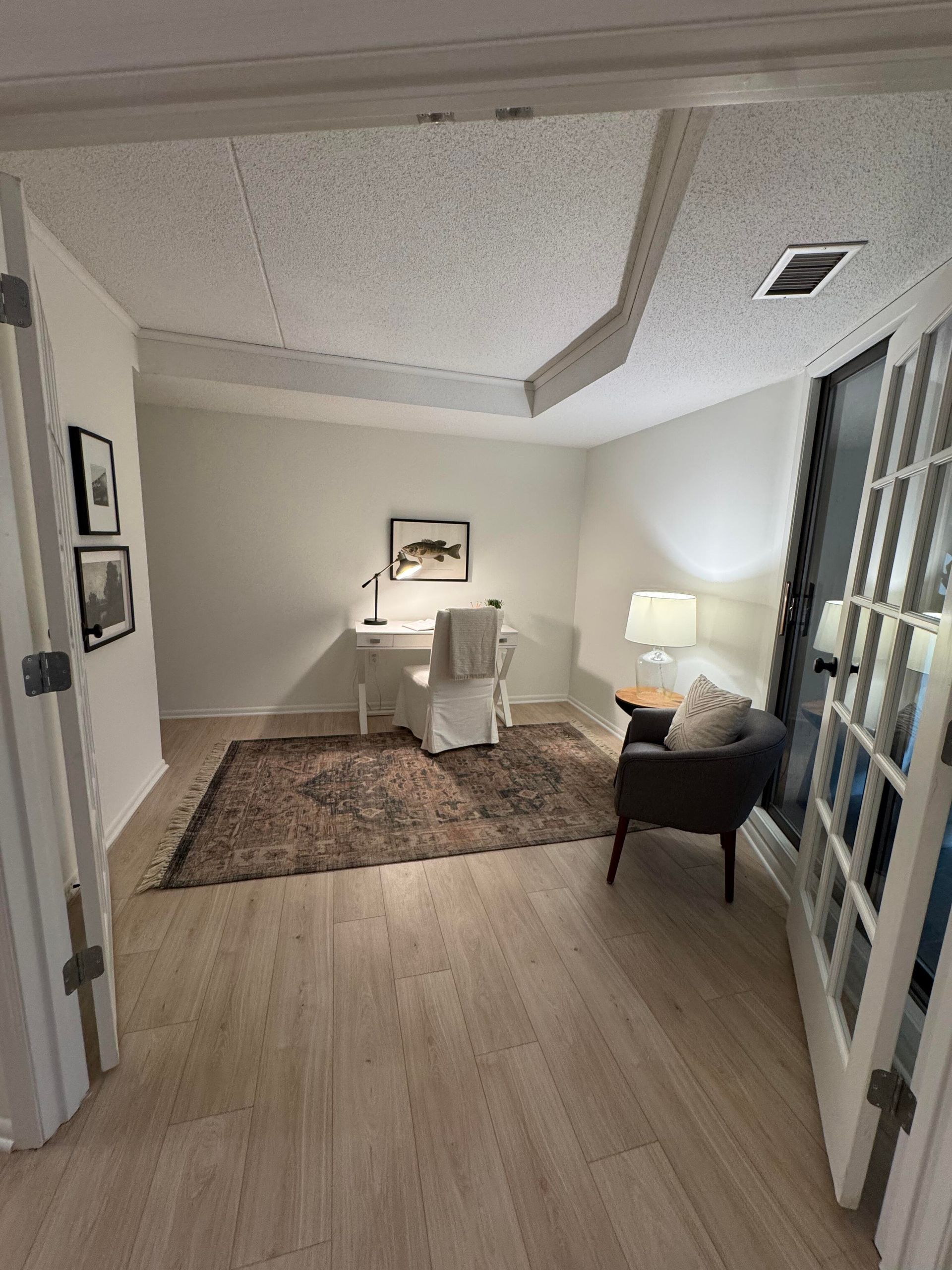 Room with desk, chair, rug, lamp, and French doors; light wood floors, white walls.