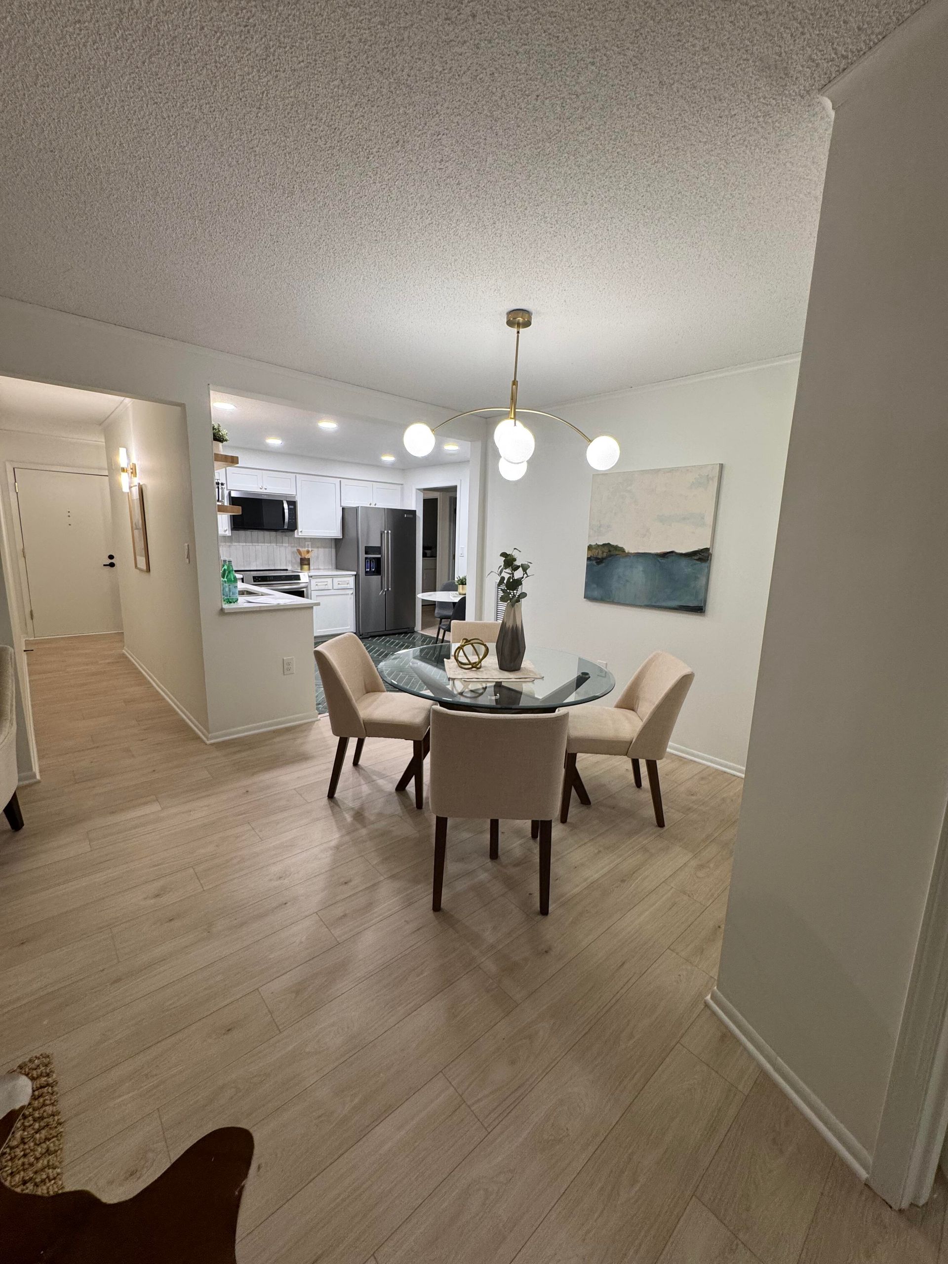 Dining area with round table and four chairs. Kitchen in background. Neutral colors, light wood floors.