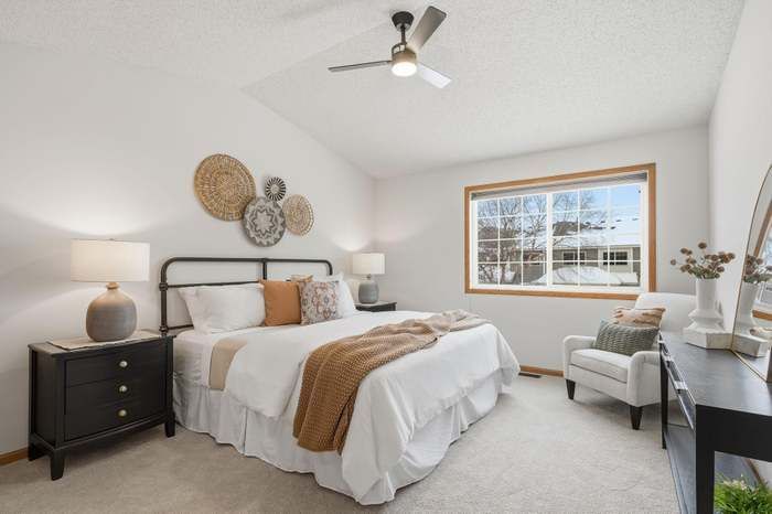Bedroom with white walls, bed with tan throw, nightstand, ceiling fan, wicker wall decor, window.
