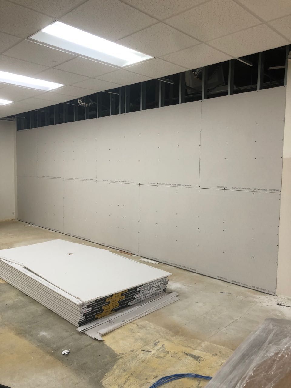 Drywall installation in progress; sheets stacked on floor. Exposed framing above wall.