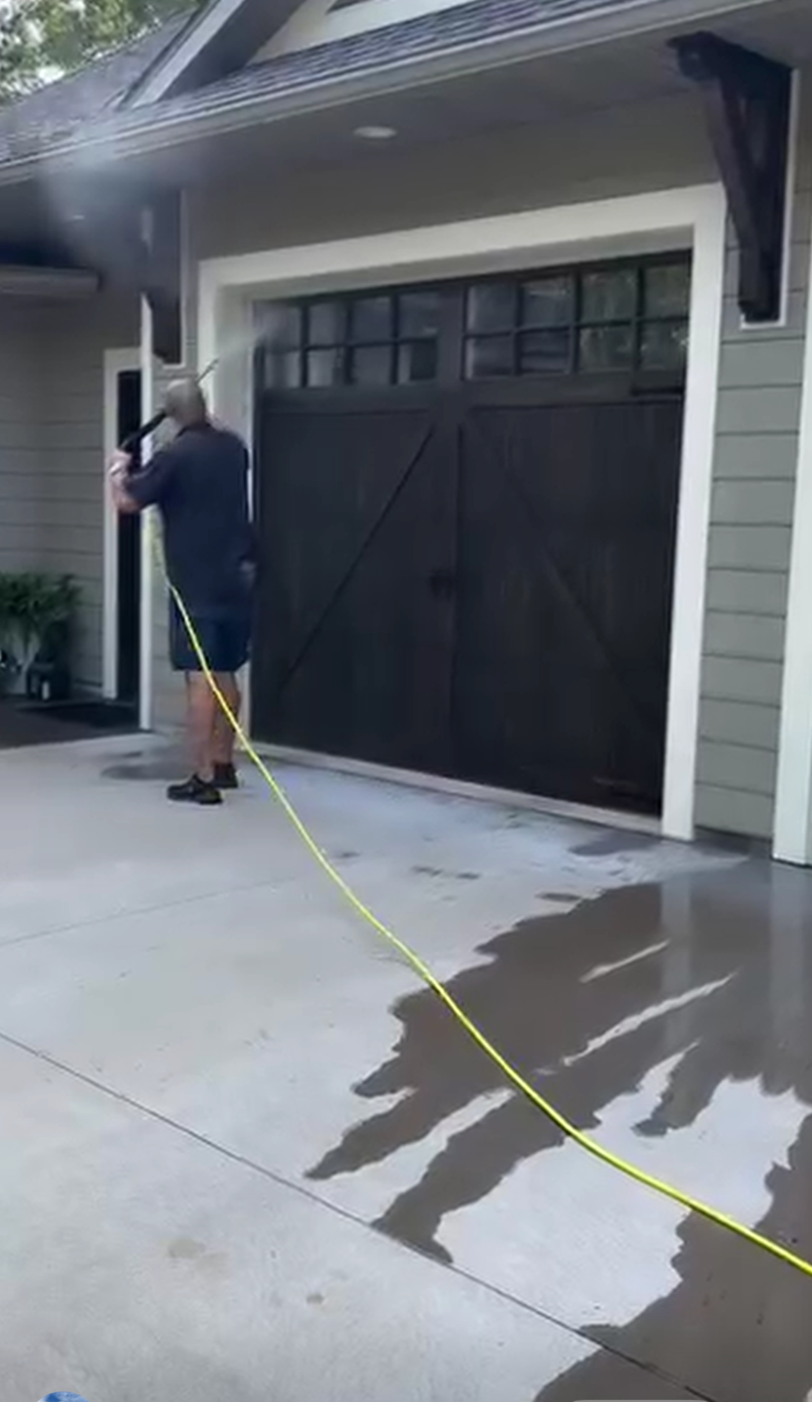 Person pressure washes a dark brown garage door, standing on a wet concrete driveway.