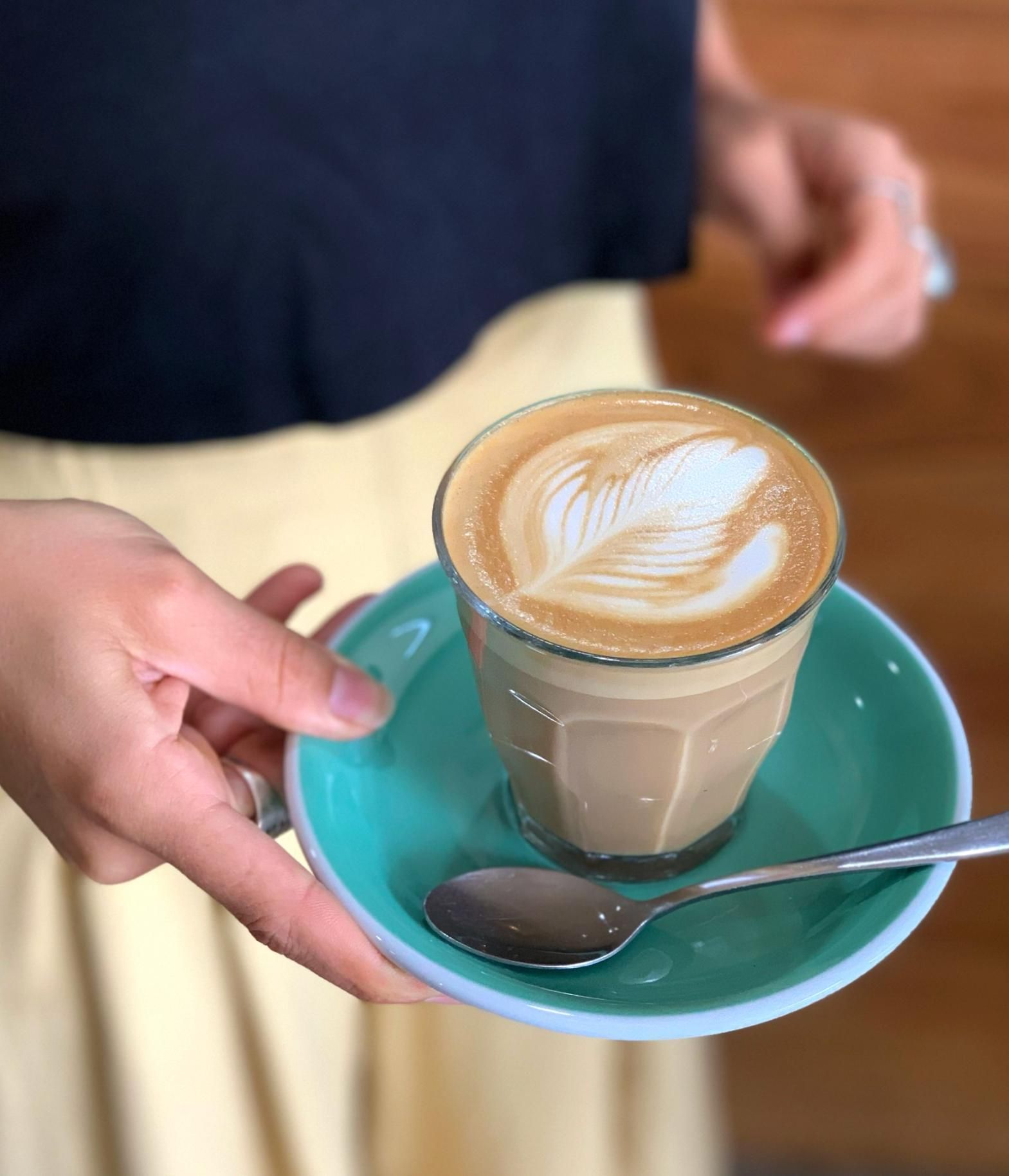 A Person Is Holding A Cup Of Coffee On A Saucer With A Spoon — The Real Grocer In Golden Beach, QLD
