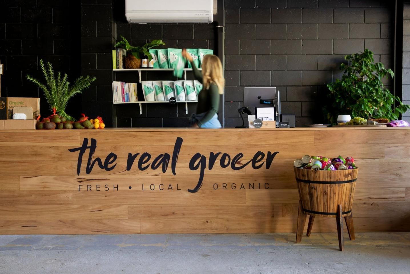 A Woman Is Standing Behind A Wooden Counter At The Real Grocer — The Real Grocer In Golden Beach, QLD