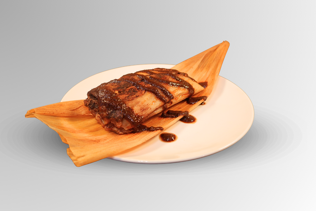 A tamale on a white plate, drizzled with dark sauce, atop a corn husk.