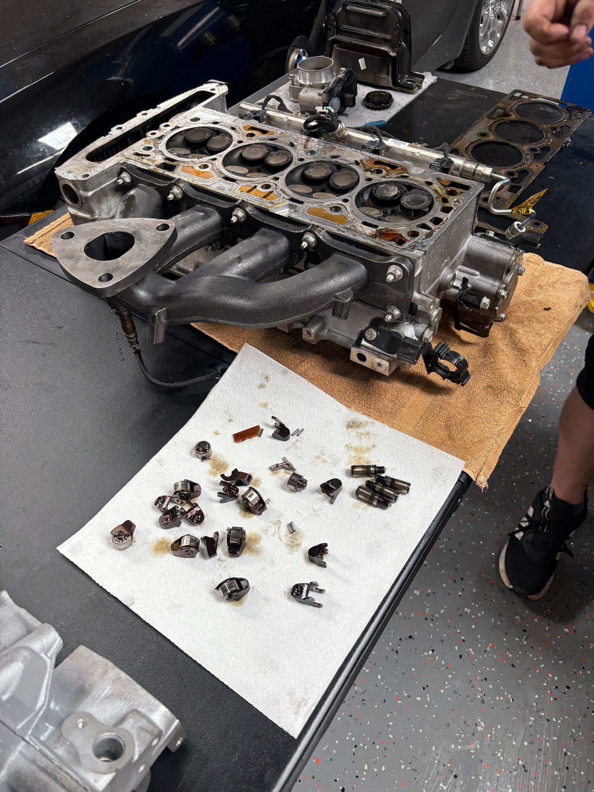 Engine head disassembled on a table with parts, a cloth, and a person's legs visible in a workshop.