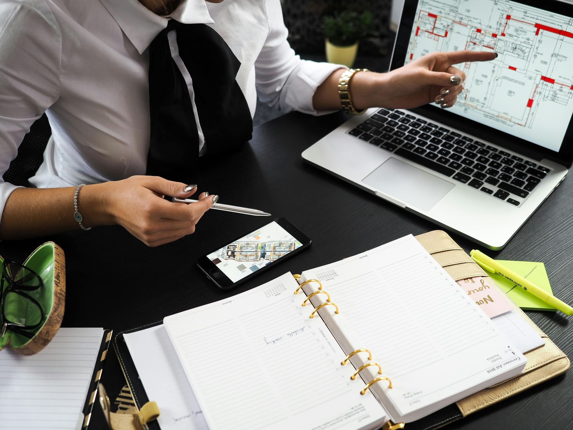 Woman pointing at floor plan on laptop; desk with planner, phone, and pen.