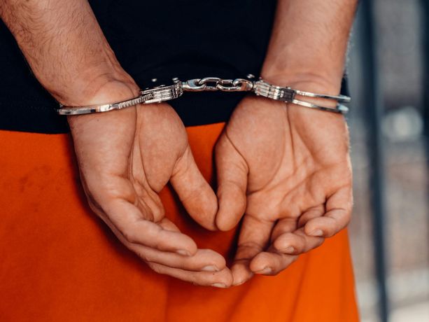 Hands cuffed behind a person wearing an orange jumpsuit and black shirt.