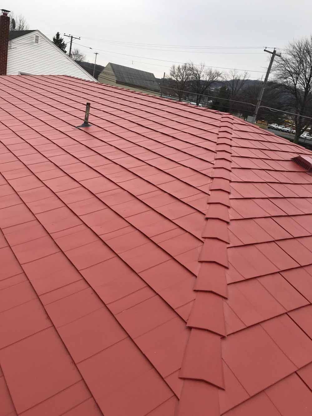 Red roof with overlapping shingles, a vent pipe, and rooftops in the background.