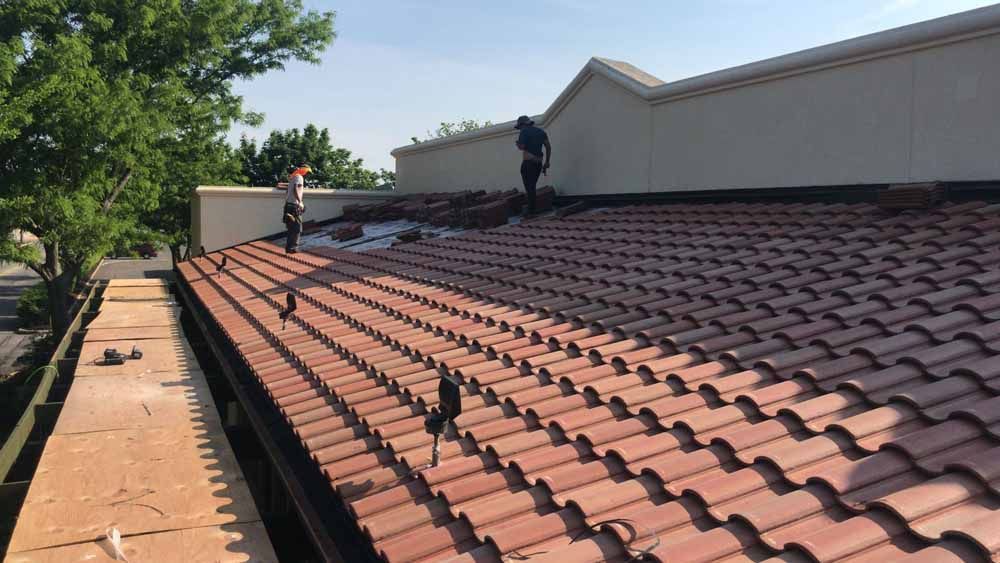 Two workers on a red tile roof, likely repairing it, with a white building and trees in the background.