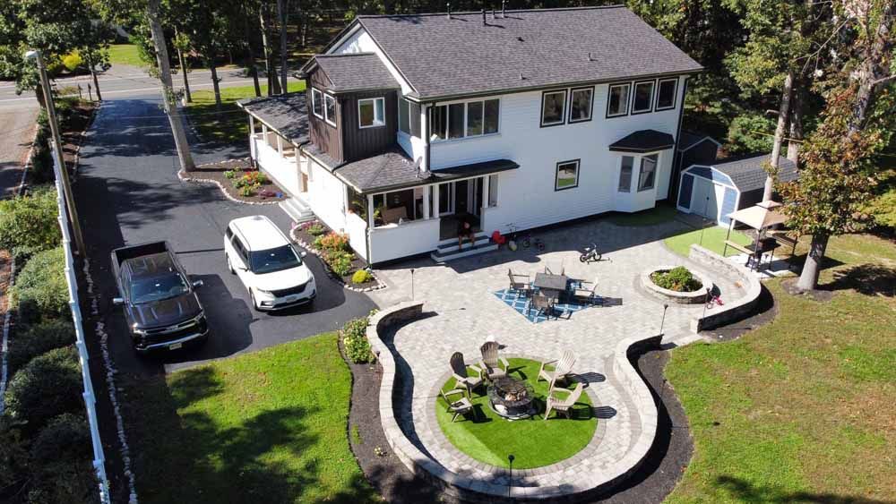Two-story house with a stone patio, fire pit, cars in driveway, surrounded by green grass and trees.