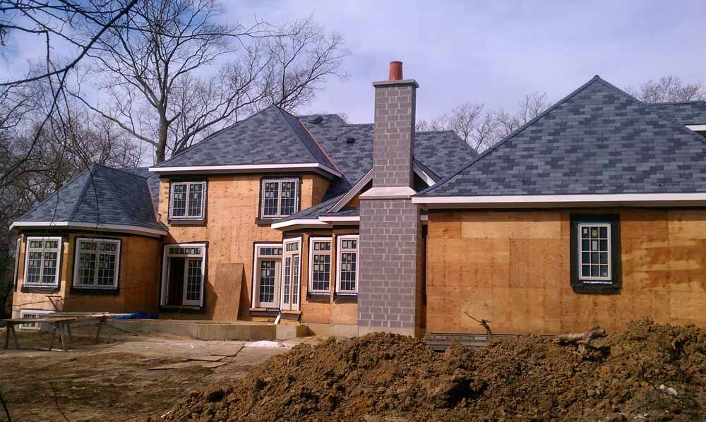 House under construction with gray shingled roof, exposed wood siding, brick chimney, and surrounding dirt.