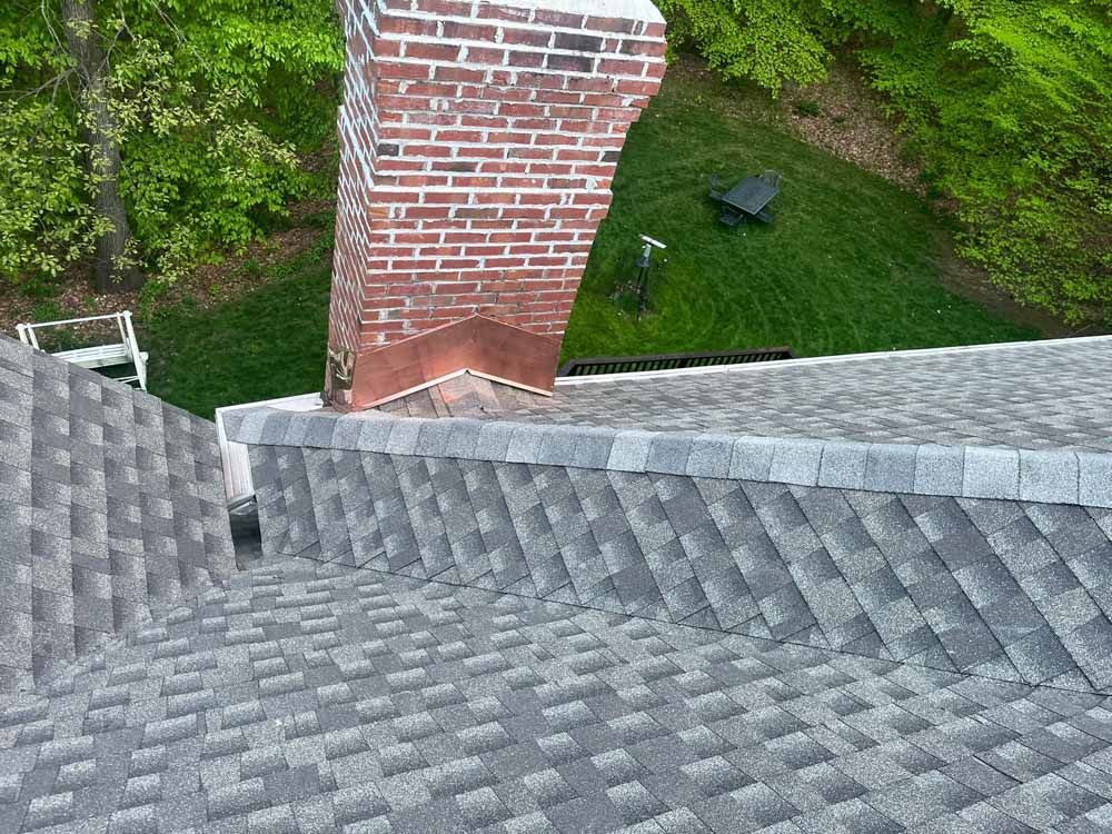 View of a roof with gray shingles, a brick chimney, and a green grassy yard.