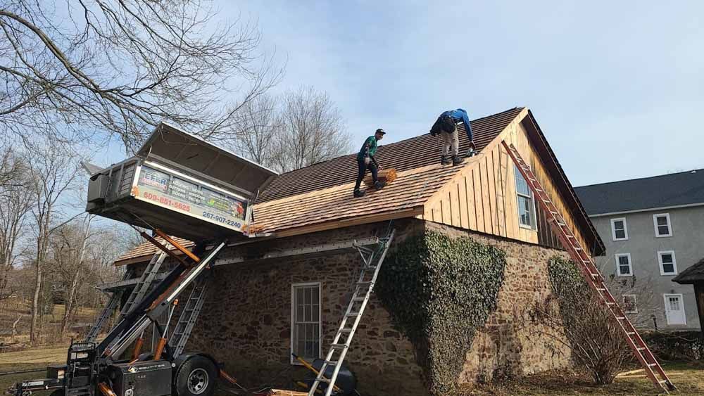 Two workers on a roof replacing tiles, with a lift platform for materials, near a brick building.
