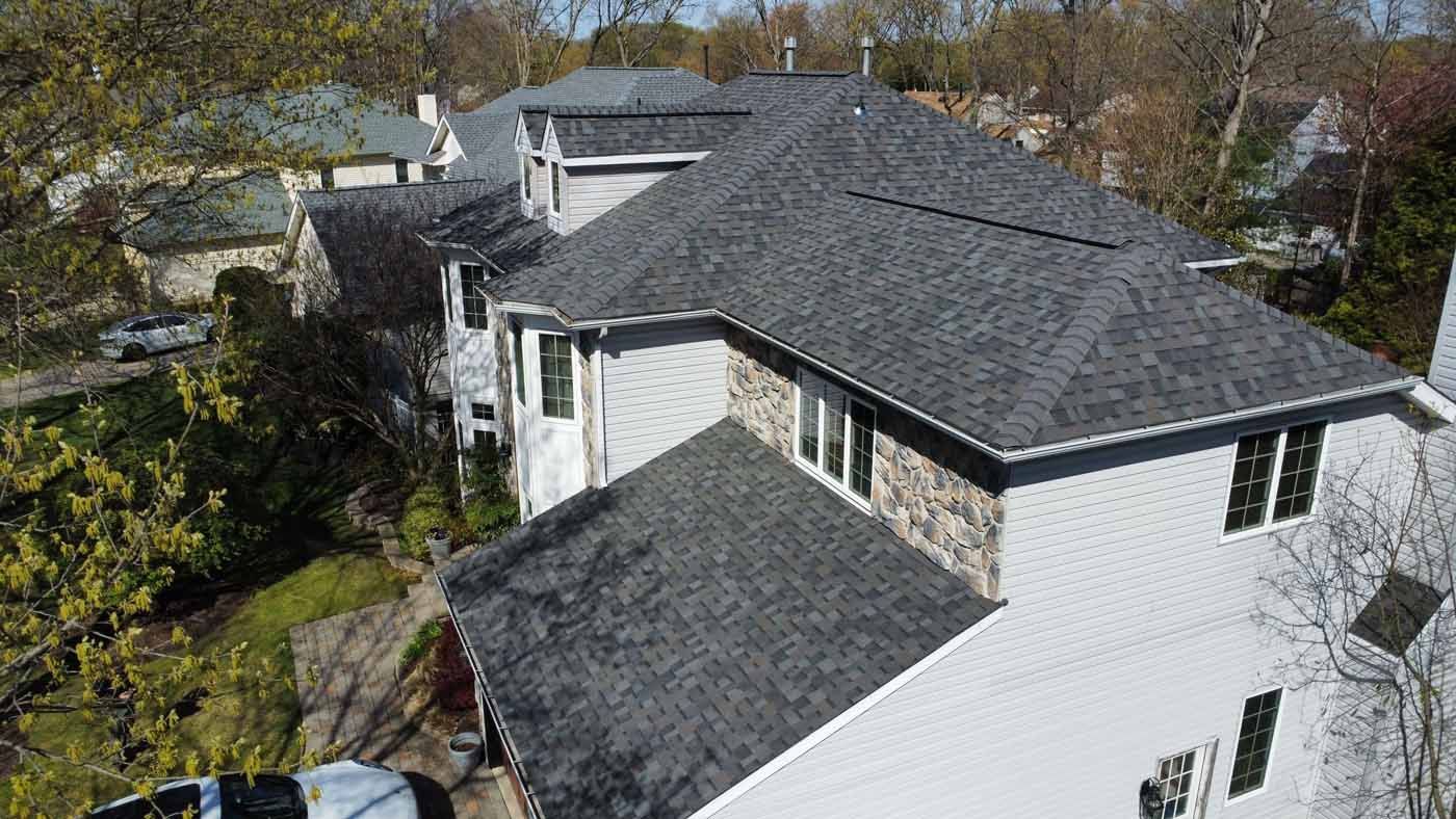 Gray-roofed house, exterior view. White siding with stone accent, windows. Trees surround the house.