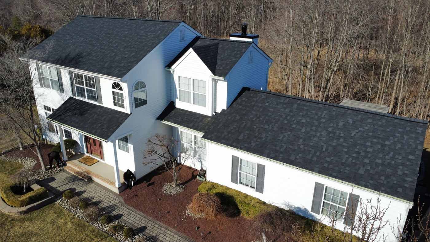 White two-story house with black roof, surrounded by brown mulch, bare trees, and a lawn on a sunny day.