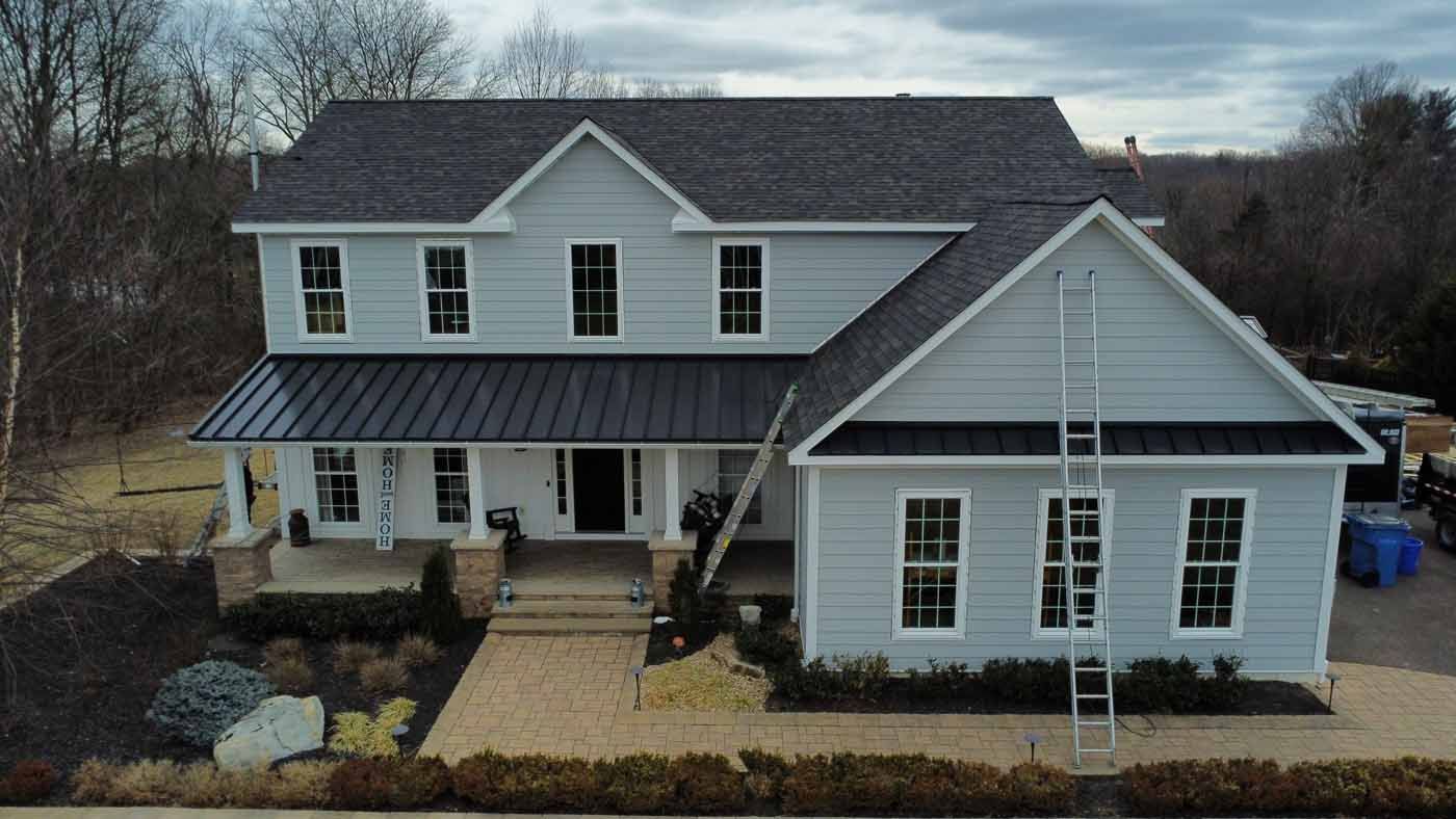 Two-story house, light blue siding, black roof and accents, covered porch, trees in background.
