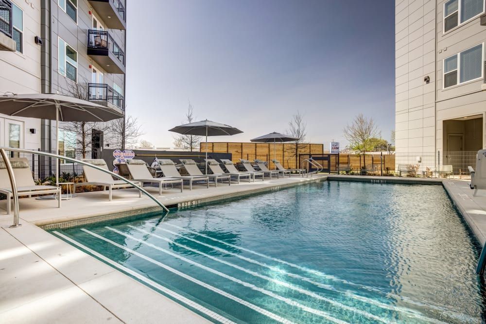 A modern apartment complex outdoor swimming pool area with loungers, umbrellas, and clear blue water on a sunny day, surrounded by the building's white balconies and a wooden privacy fence at 44 South in Austin, TX.