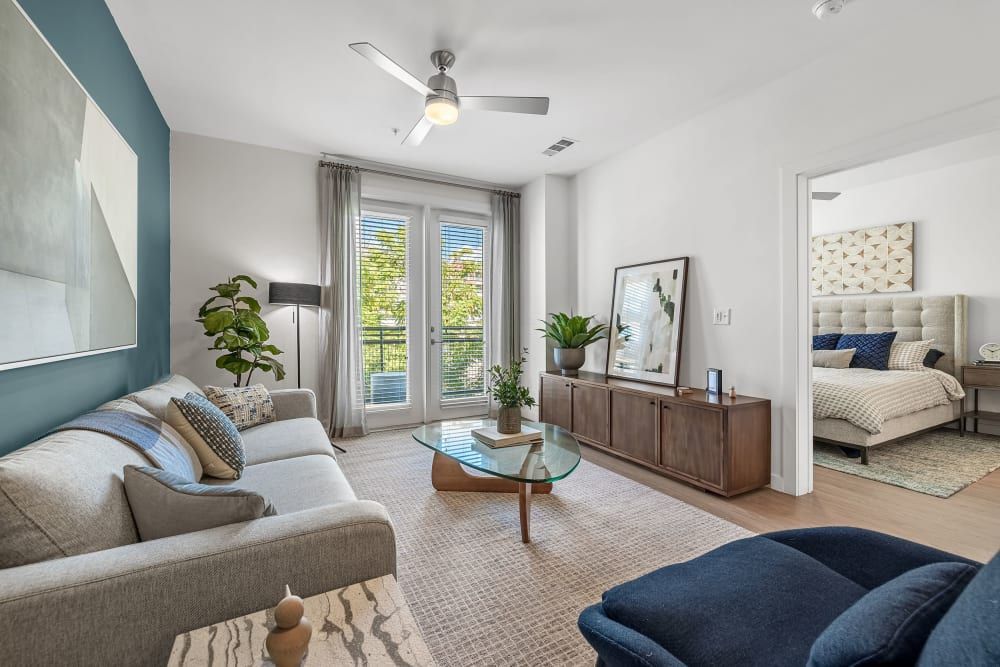 Modern living room interior in neutral tones with a beige sofa, blue accent chairs, and a mid-century style wooden sideboard. A large abstract painting hangs on the accent wall, and through the glass door, a balcony with greenery is visible. The room opens to a cozy bedroom with a tufted headboard at 44 South in Austin, TX.