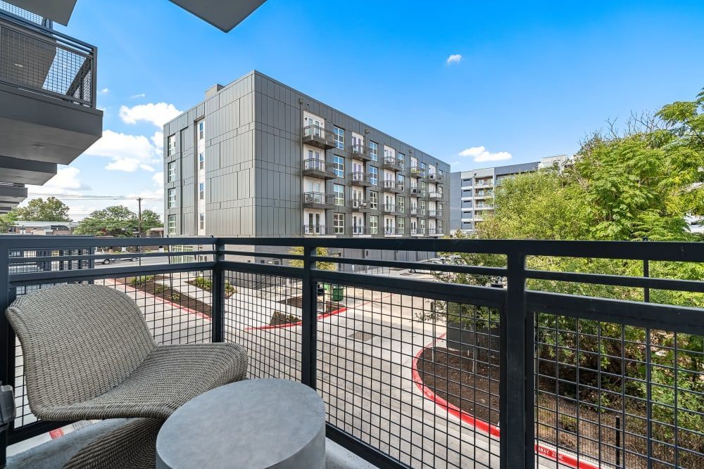 View from a balcony featuring a single woven chair and a small table, overlooking a modern apartment building with a clear sky above. Green trees are visible on the right, with a glimpse of the parking area below at 44 South in Austin, TX.