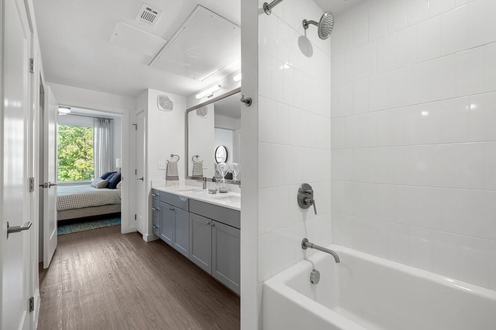 Modern bathroom interior with a view into a bedroom. The bathroom features a vanity with blue cabinets and double sinks, large mirrors, and white subway tiles. A bathtub with a shower head is visible to the right, reflecting a clean and bright design. The adjacent bedroom has a bed with blue bedding and a window showing green foliage outside at 44 South in Austin, TX.