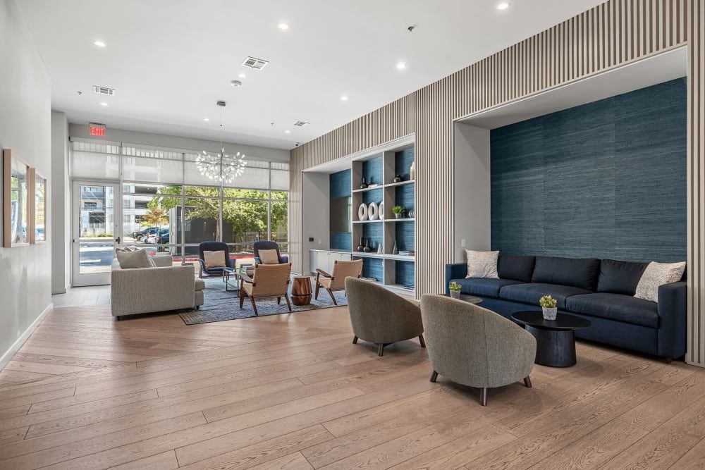 Modern clubhouse area with herringbone wood flooring, a blue sectional sofa, two gray armchairs, wooden accent chairs, and a bookshelf. A large front window lets in natural light, and a stylish chandelier hangs from the ceiling. The color scheme is neutral with blue accents at 44 South in Austin, TX.