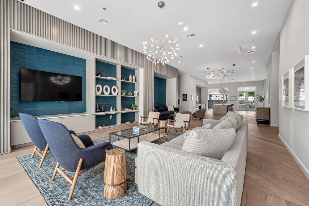 A modern living room area featuring a long, creamy sofa, two blue armchairs on a blue rug, and a sleek built-in wall unit with a flat-screen TV and decorative shelves against a vibrant blue backdrop. The room is bright with natural and artificial lighting, showcasing a combination of wood floors, white walls and ceiling, and elegant hanging light fixtures at 44 South in Austin, TX.