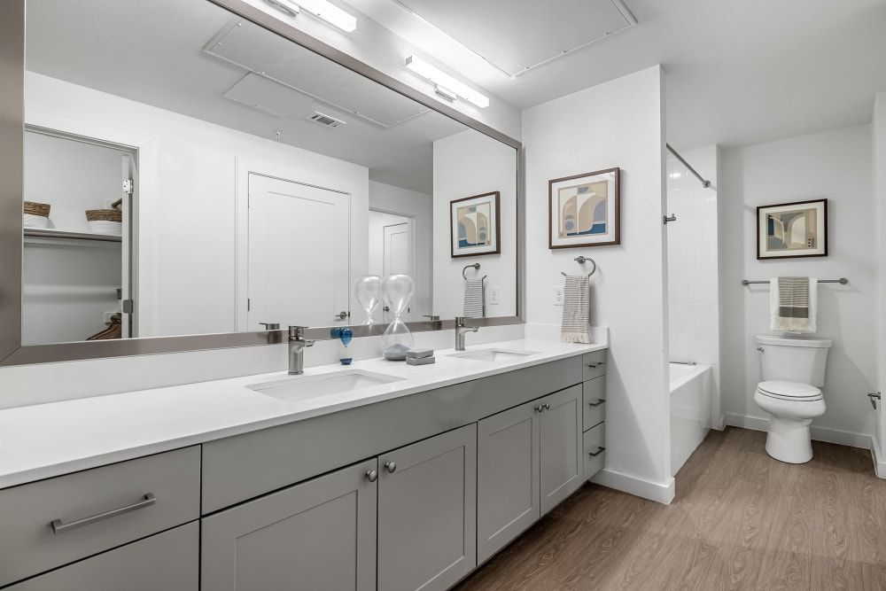 Modern bathroom featuring a long vanity with dual sinks, large mirrors, and white countertops against gray cabinets. There are decorative elements such as framed abstract artwork on the walls and hourglass on the counter. The room includes overhead recessed lighting, a shelved nook, a towel bar with striped towels, and a shower with a clear glass door in the background, near a standard toilet. The floor is covered in vinyl flooring that mimics wood texture at 44 South in Austin, TX.