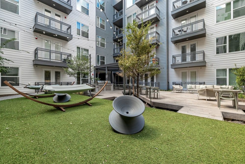 A modern apartment building courtyard featuring amenities such as a green artificial turf area, a wooden deck with outdoor seating arrangements, hammocks, and sculptural decorative elements. Trees are strategically placed around the space, and multiple balconies overlook the communal leisure area at 44 South in Austin, TX.
