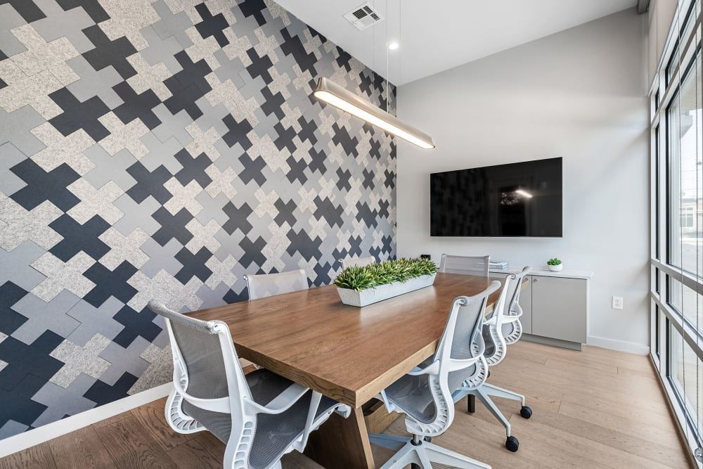 Modern office meeting room with a wooden table and white rolling chairs, geometric pattern wall, hanging linear light, and a large window providing natural light. A flat-screen monitor is mounted on the opposite wall, and indoor plants add a touch of greenery at 44 South in Austin, TX.