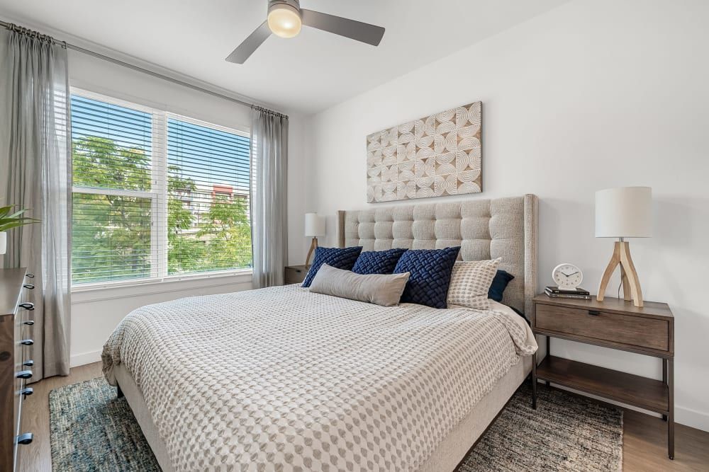 Modern bedroom interior featuring a large bed with a tufted headboard, geometric-patterned bedding, and multiple pillows. A wooden nightstand holds a lamp and an alarm clock. Sheer curtains soften the natural light from a window with a view of green foliage. A decorative wall panel hangs above the bed, and a ceiling fan is mounted above at 44 South in Austin, TX.