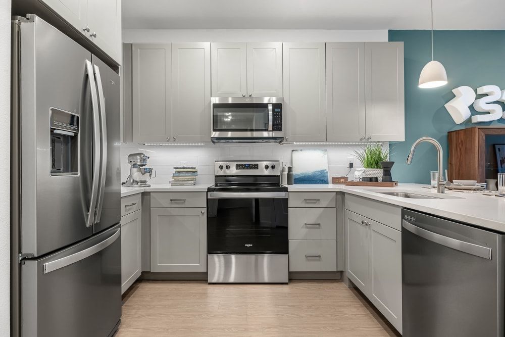 Modern kitchen interior with stainless steel appliances, light grey cabinets, and a teal accent wall. There is a standing refrigerator, a microwave over a range, and a dishwasher. The kitchen includes a light wood floor, white countertops, and a hanging white lamp. Decorative touches include a stand mixer, plants, and neatly arranged books and kitchenware at 44 South in Austin, TX.