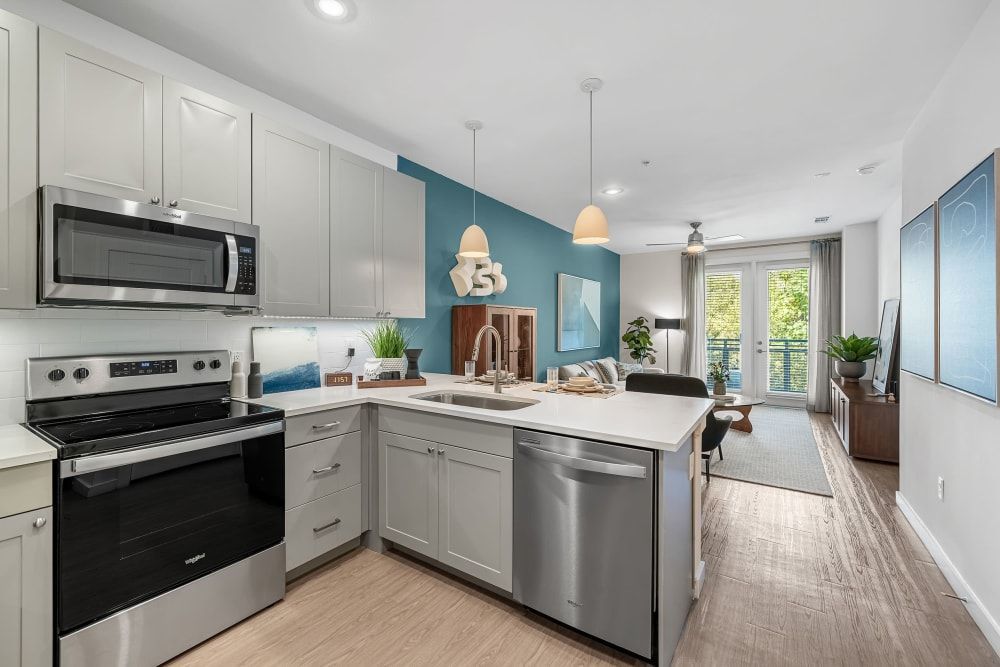 Modern kitchen with white cabinetry, stainless steel appliances, and an island extending into a cozy living area with a sofa, chairs, and a television. The space is brightly lit with natural light from a balcony door and features a combination of soft teal and white walls, accented with framed wall art at 44 South in Austin, TX.