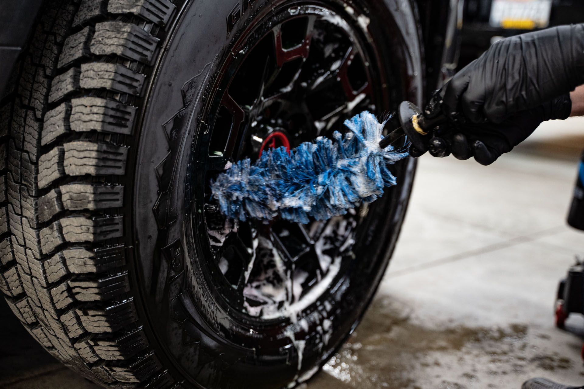 A gloved hand uses a blue, soapy microfiber brush to scrub a black car wheel.