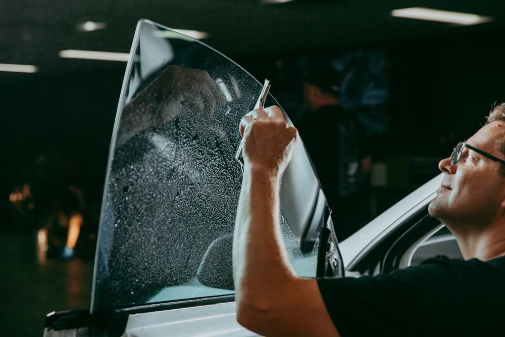 A professional applies window tint to a car door glass, smoothing the wet film with a small tool in a workshop.