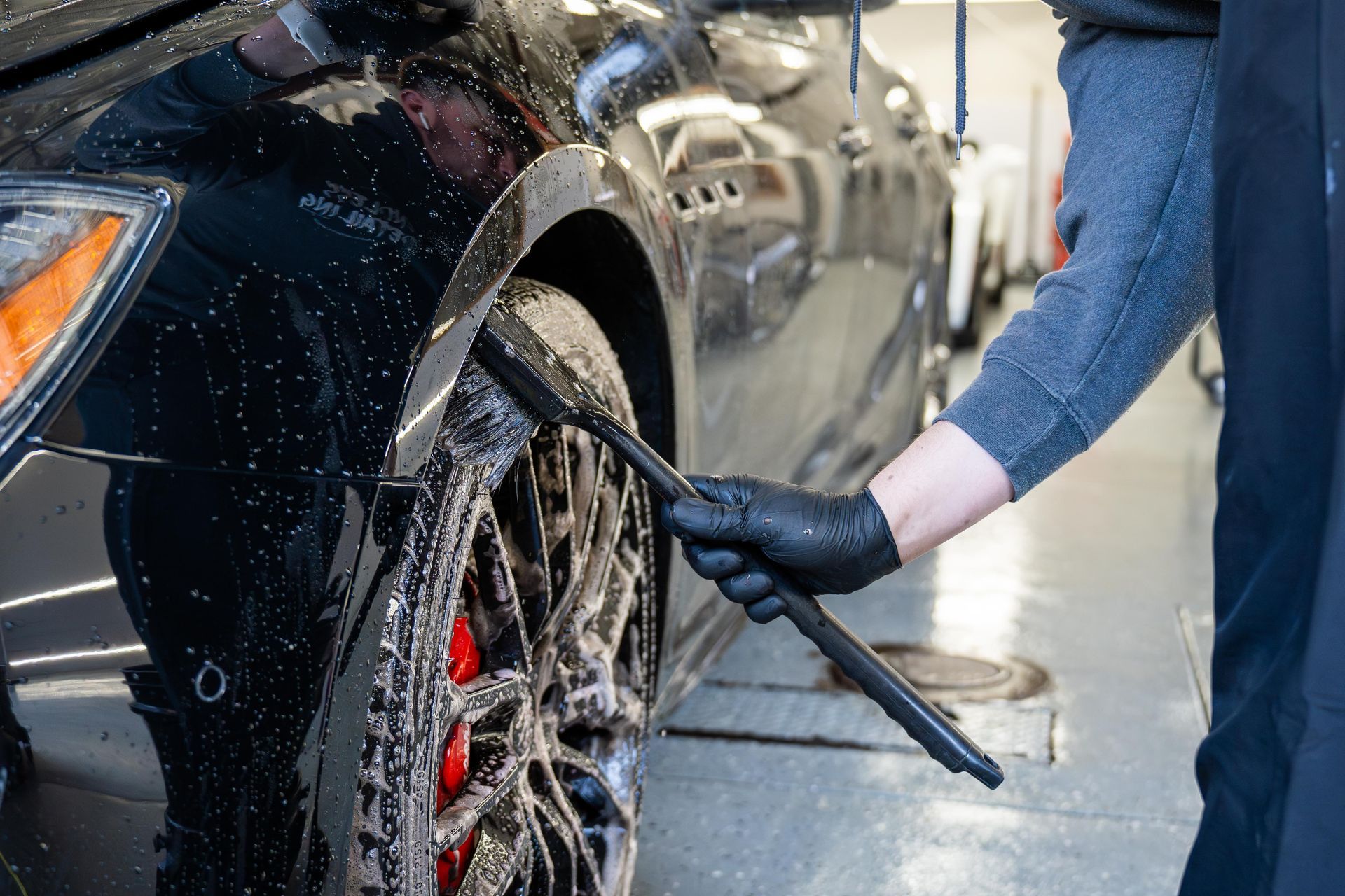A person wearing black gloves uses a long-handled brush to clean a soapy car wheel at a shop.