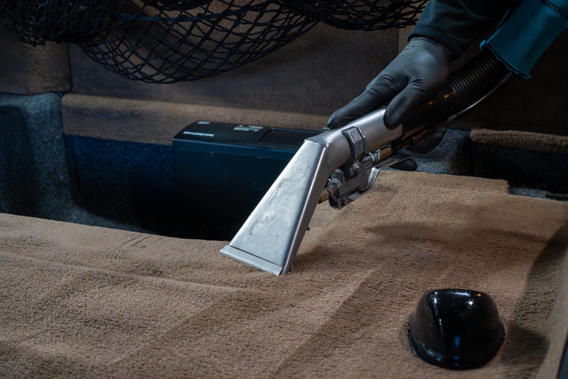 A gloved hand uses a vacuum extraction tool to deep clean the tan fabric upholstery of a vehicle interior.