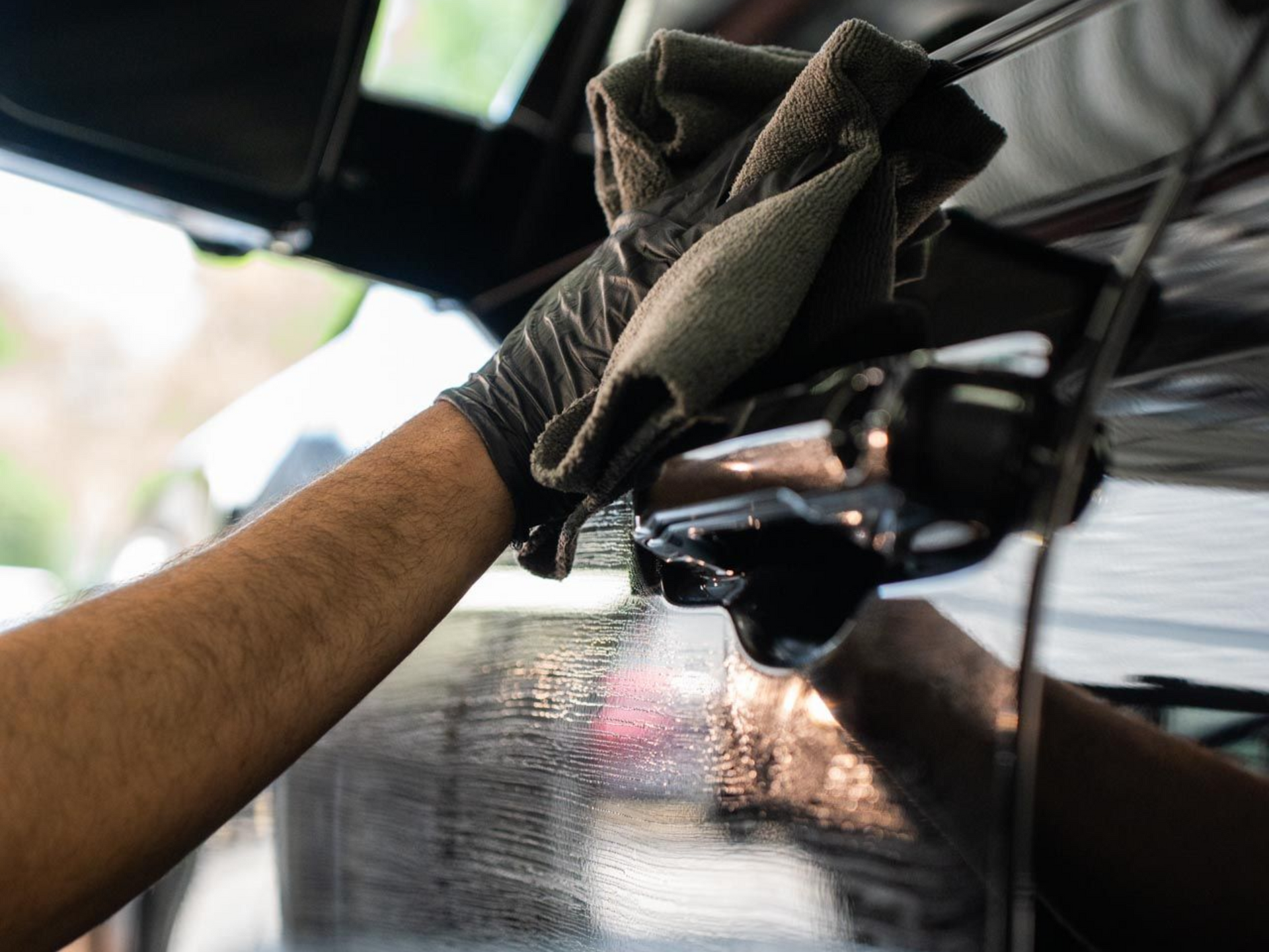 A hand wearing a black glove uses a cloth to wipe down the reflective black door of a vehicle.