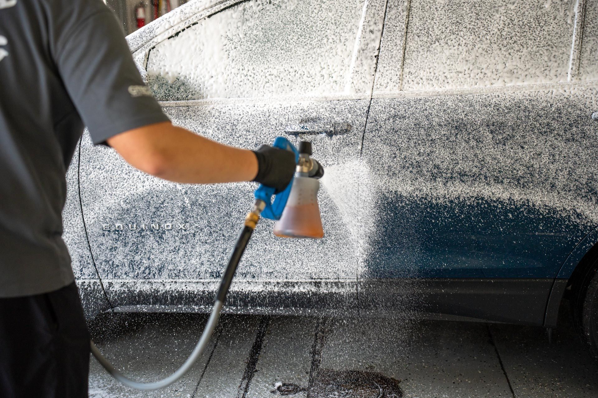 A worker in a gray shirt uses a spray nozzle to apply white cleaning foam to the side of a car in a service bay.