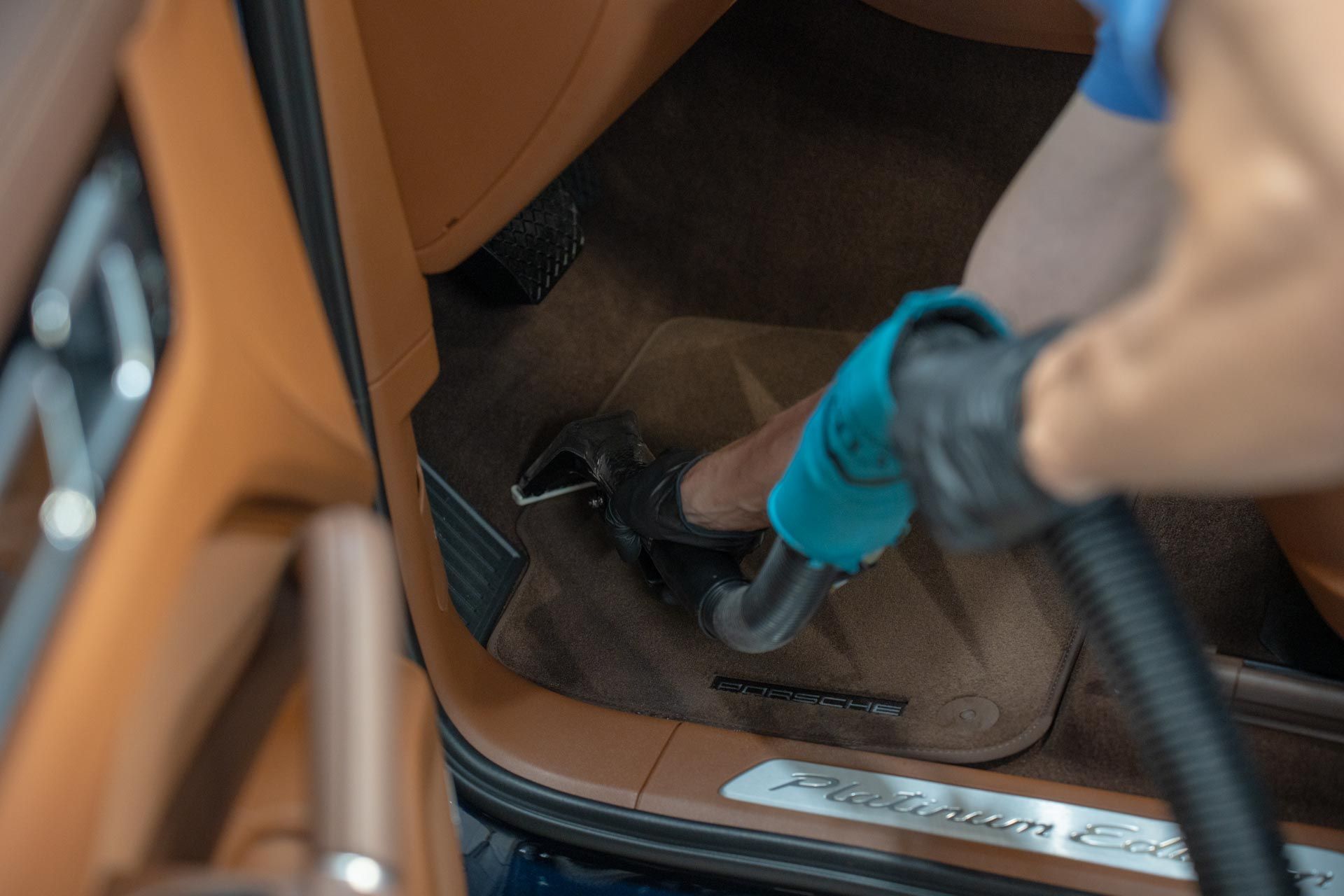 A person wearing blue gloves vacuums the brown carpeted floor of a luxury car interior.