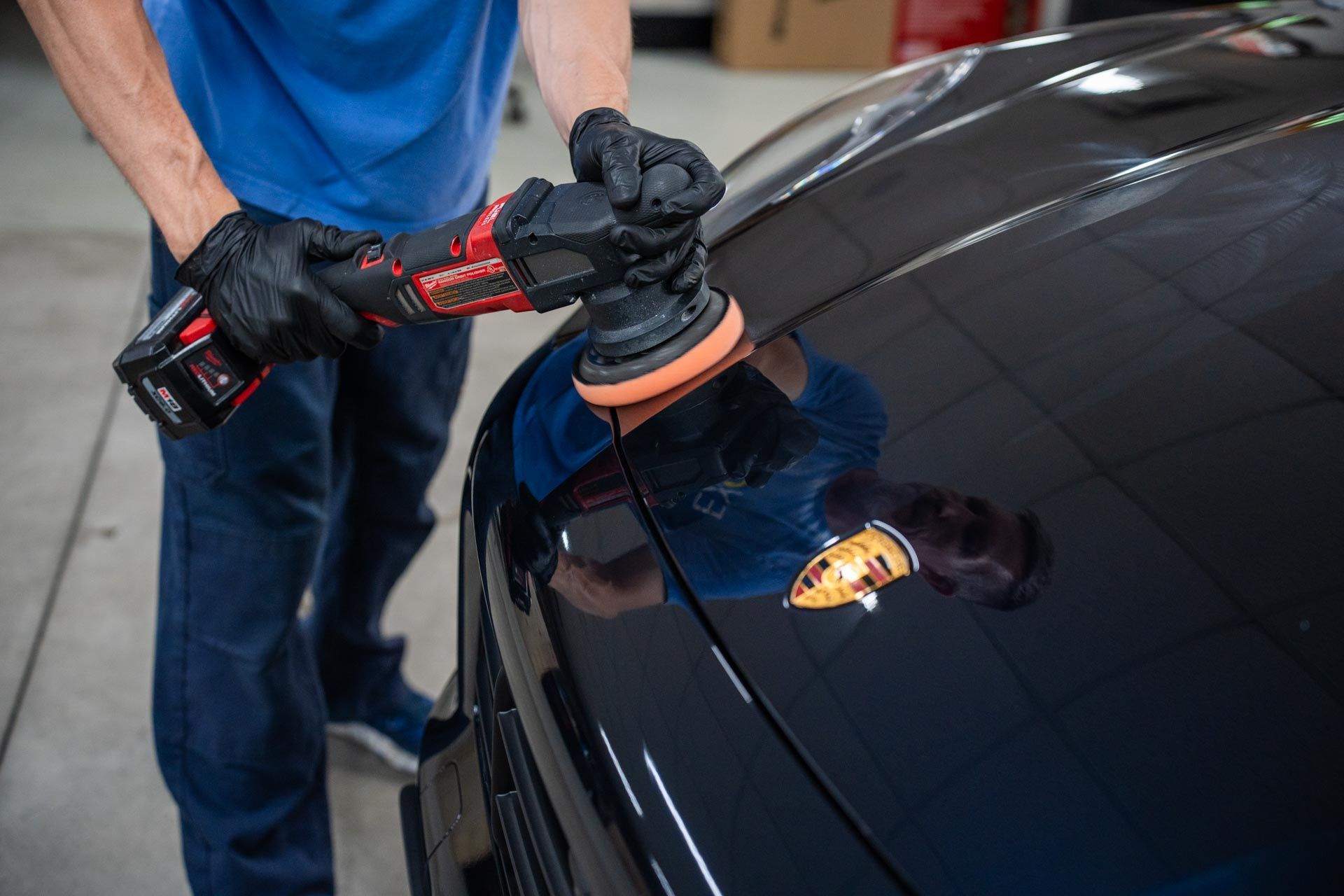 A technician in black gloves uses a red power polisher to detail the hood of a black Porsche.