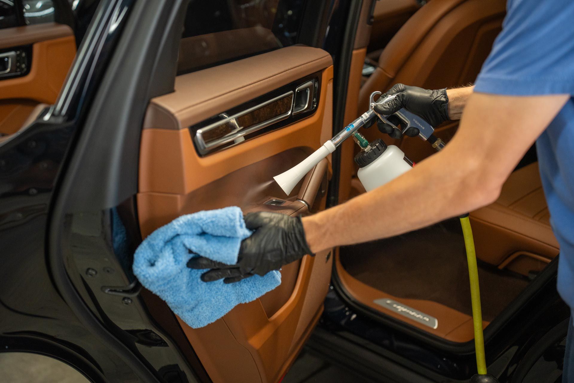 A worker in black gloves uses a pneumatic cleaning tool and a blue microfiber cloth to clean a brown leather car door.