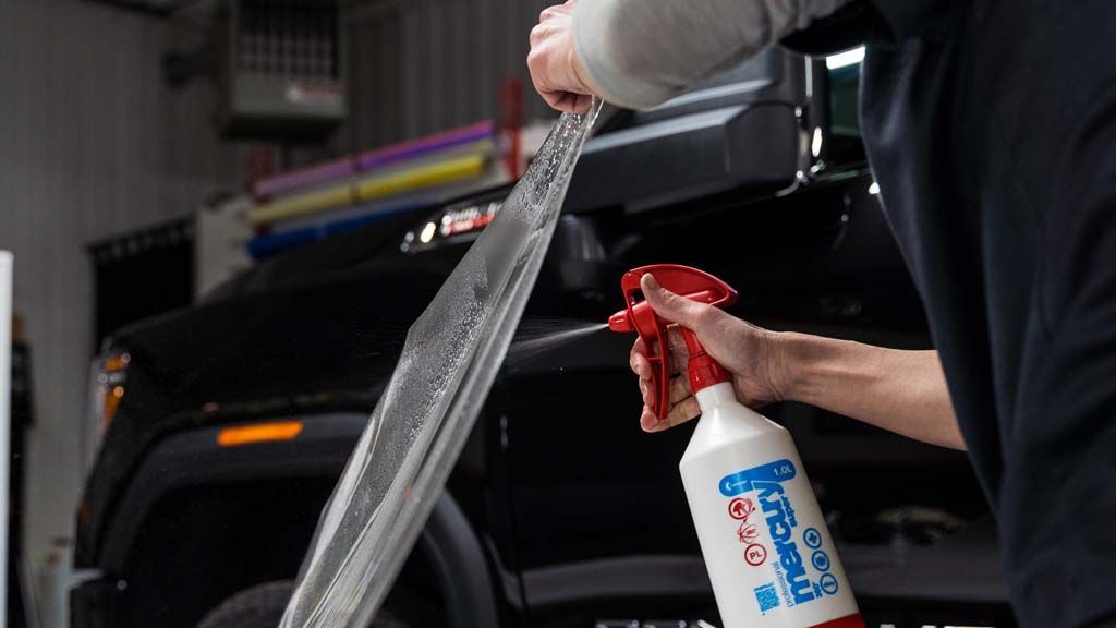 A person sprays liquid from a red and white spray bottle onto a piece of transparent adhesive film held near a black truck.