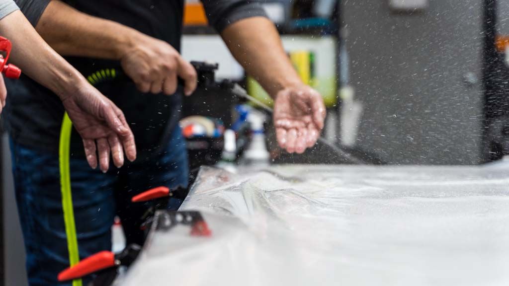 Two people use a spray nozzle to apply water to a large, clear sheet of material secured with red clamps on a table.