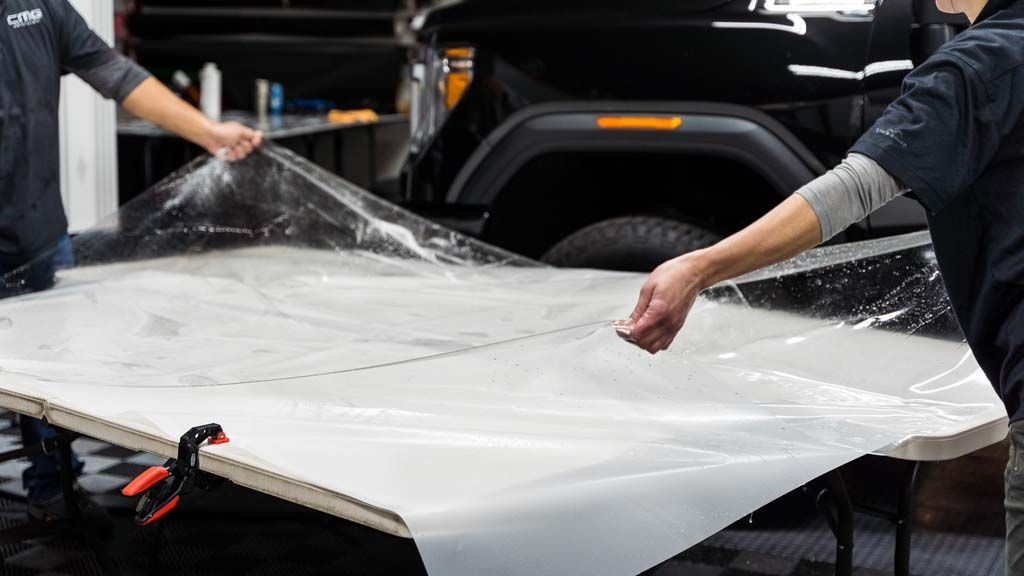 Two people work together to carefully smooth a sheet of clear protective film onto a large table in an automotive shop.